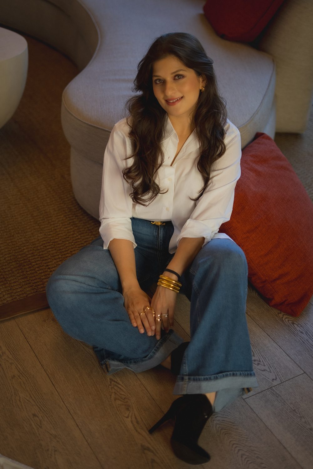 Portrait of Devika Apte sitting on a wooden floor in a white shirt and jeans, smiling